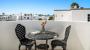 A round table with two chairs, a book, and a vase with flowers on a balcony overlooking a residential area with white buildings and palm trees. - Villa Sheila – Family Villa with Heated Pool in Puerto del Carmen (Puerto del Carmen)