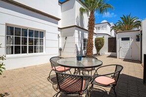 A patio with a round table and wicker chairs, a palm tree, and a white building with large windows.