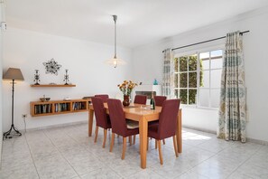 A dining area with a wooden table and chairs, a hanging light fixture, a bookshelf, and a window with curtains.