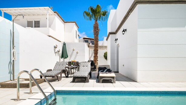 A swimming pool with a tiled edge, lounge chairs, a palm tree, and a white building with a balcony. - Villa Sheila – Family Villa with Heated Pool in Puerto del Carmen (Puerto del Carmen)