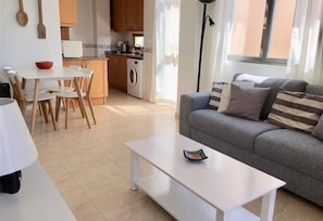 Un salon moderne avec un canapé gris, un coin repas avec une table et des chaises, une cuisine équipée d’un lave-linge et une lampe sur pied.