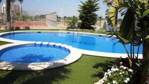 A swimming pool with a surrounding deck, palm trees, and a tennis court in the background. - Cosy Bright & Neat Apartment with Sea View, Shared Pool & Wi-Fi (Guardamar del Segura)