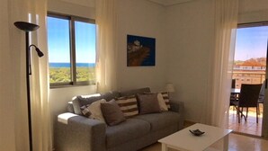 A living room with a sofa, a coffee table, a lamp, and a view of the sea through a window. - Cosy Bright & Neat Apartment with Sea View, Shared Pool & Wi-Fi (Guardamar del Segura)