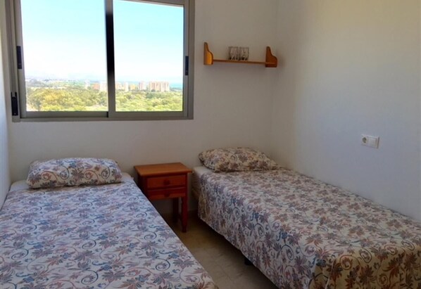 Ein Schlafzimmer mit zwei Betten, einem kleinen Holfnachtkästchen, einem Regal mit Büchern und einem Fenster mit Blick auf die Stadt.