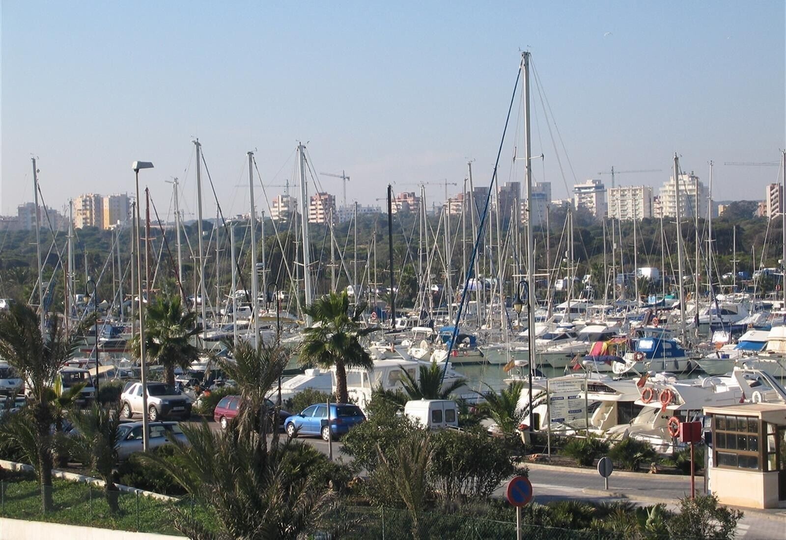 A marina with numerous boats docked, surrounded by palm trees and a city skyline in the background.