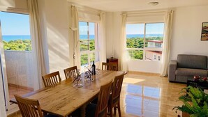 Un comedor con mesa y sillas de madera, ventanas grandes con cortinas, un sofá y una planta en maceta.