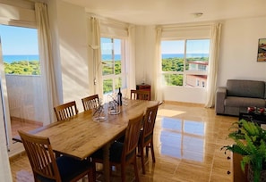 Un comedor con mesa y sillas de madera, ventanas grandes con cortinas, un sofá y una planta en maceta.
