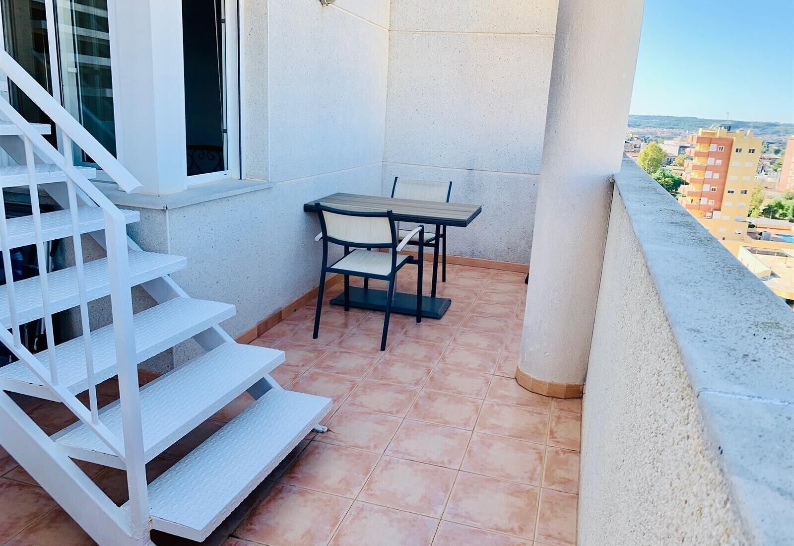 Un balcón con una mesa y sillas, una escalera y vistas a edificios a lo lejos.