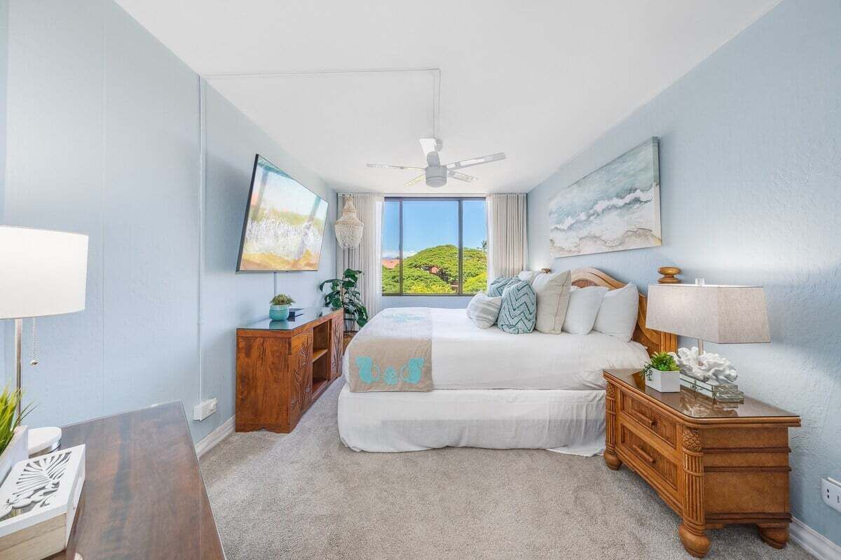 A bedroom with a bed, a nightstand, a TV, and a window with a view of greenery.