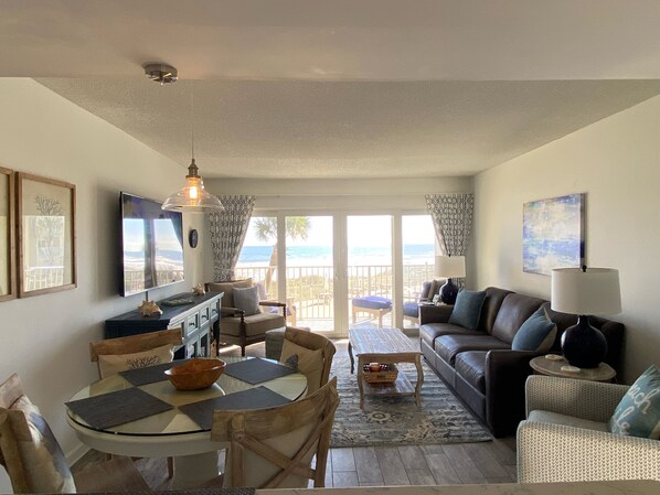 Un salon comprenant un coin repas, un canapé, une table basse et un téléviseur. On peut admirer la vue sur la plage à travers les portes vitrées.