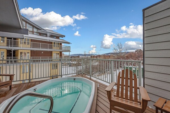 A deck with a hot tub, wooden chairs, and a view of a multi-story building with balconies.