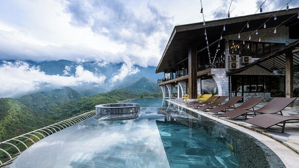 Una piscina infinita con vistas a montañas, sillones de descanso y un área de restaurante con mesas al aire libre.