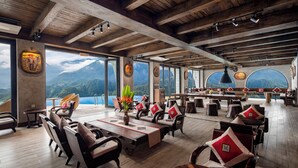 Une salle à manger spacieuse, dotée de tables et de chaises en bois, de grandes fenêtres offrant une vue sur les montagnes et d’un plafond haut avec des poutres apparentes.