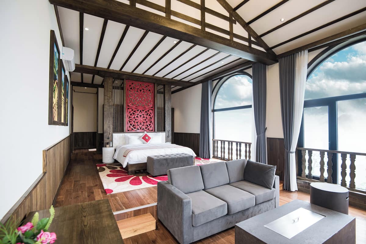 Una habitación de hotel moderna con una cama grande, un sofá gris, una mesita pequeña y un balcón con vistas al cielo.