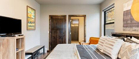 A bedroom with a bed, a TV, a bench, and a door leading to another room.