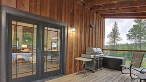 A wooden deck with a grill and chairs, a glass door leading to an interior with a sofa and table, and a view of trees. - Dream Catcher- Spacious Rooms & Views- Sleeps 18 (North Lawrence)