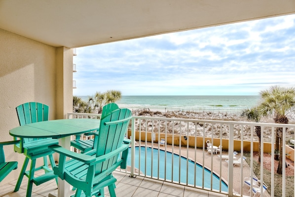 A balcony with a view of the beach, a pool, and outdoor seating.