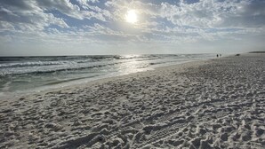 Una playa arenosa con huellas y olas, bajo un cielo parcialmente nublado con el sol brillando.