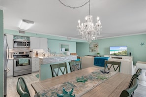 A modern kitchen with stainless steel appliances, a dining area with a wooden table and chairs, and a living area with a television and artwork on the wall.