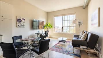 A modern living room with a sofa, a coffee table, and a dining area with chairs and a glass table. There is a large window providing natural light and a view outside.