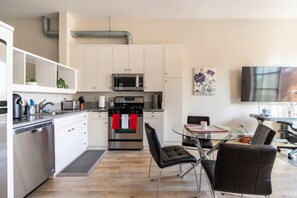 Una cucina moderna con mobili bianchi, elettrodomestici in acciaio inossidabile, lavastoviglie e una zona pranzo con tavolo in vetro e sedie nere.