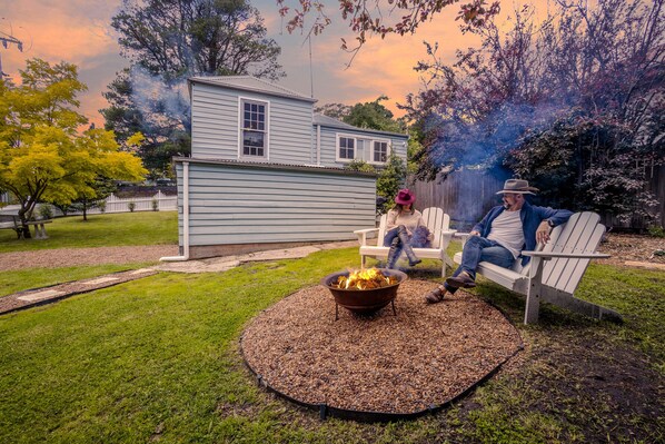 A backyard with a fire pit, a house, and a person sitting on a bench.