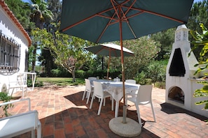 A patio with a table and chairs under a large umbrella, surrounded by greenery and a white building with a chimney. - Villa in Almancil with Pool and Golf Green (Almancil)