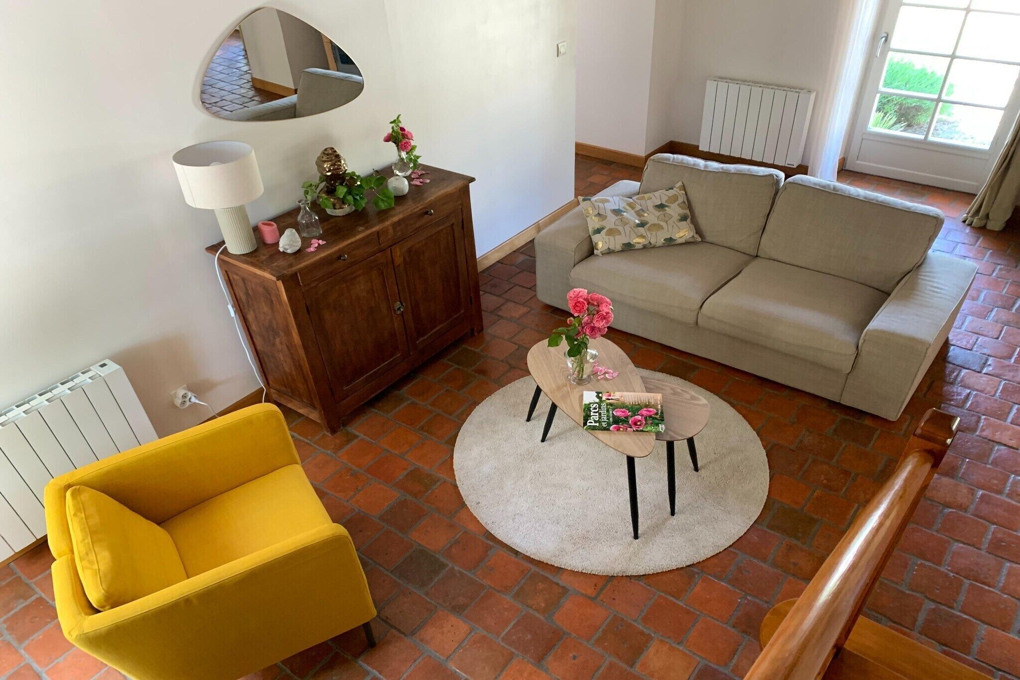 Una sala de estar con un sillón amarillo, un sofá beige, un aparador de madera y una mesa de centro redonda con flores.