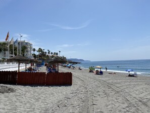Ein Strand mit Sonnenschirmen, Liegestühlen und einem Gebäude mit Balkonen.