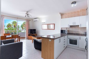 A modern kitchen with a central island, a dining area with a TV, and a balcony with a view of palm trees.