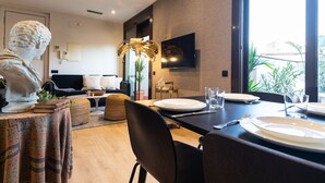 Un comedor con una mesa dispuesta para dos personas, un sofá, un televisor y un amplio ventanal con plantas afuera.