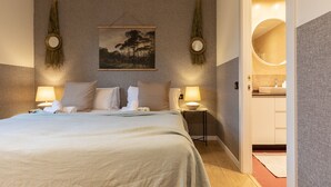Un dormitorio con cama, dos lámparas de noche, un cuadro y un baño visible a través de una puerta abierta.