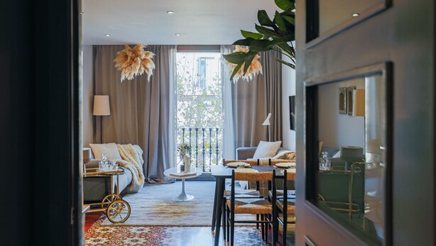 Una habitación de hotel moderna con una cama, una zona de estar con un sofá y sillas, una mesa de comedor y un gran ventanal con cortinas.