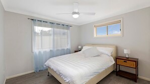 Un dormitorio con cama, mesita de noche, lámpara, ventilador de techo, ventana y cortinas traslúcidas.