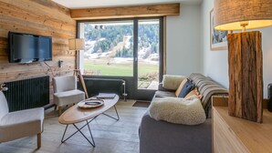 Een moderne woonkamer met een televisie, een houten salontafel, een bank en een groot raam met uitzicht op een bergachtig landschap.