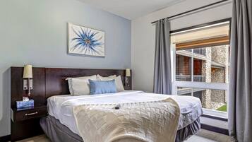 Ein Hotelzimmer mit einem großen Bett, einem Nachttisch mit Lampe, einem gerahmten Kunstwerk an der Wand und Blick auf ein Steinhaus durch ein Fenster.