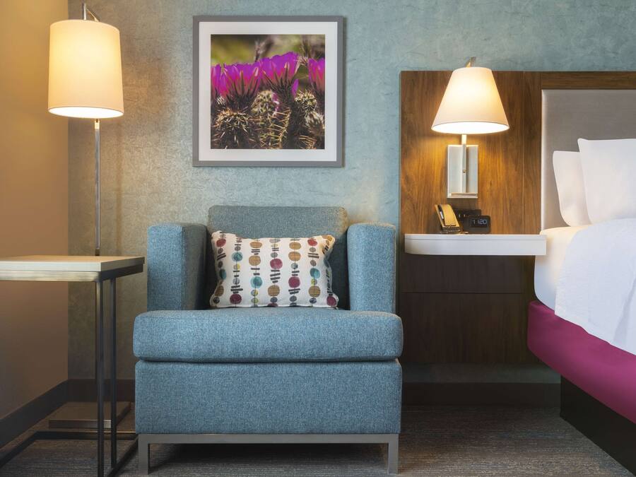 A hotel room with a blue armchair, a small table, a bed with white bedding, a nightstand with a lamp, and a framed picture on the wall.