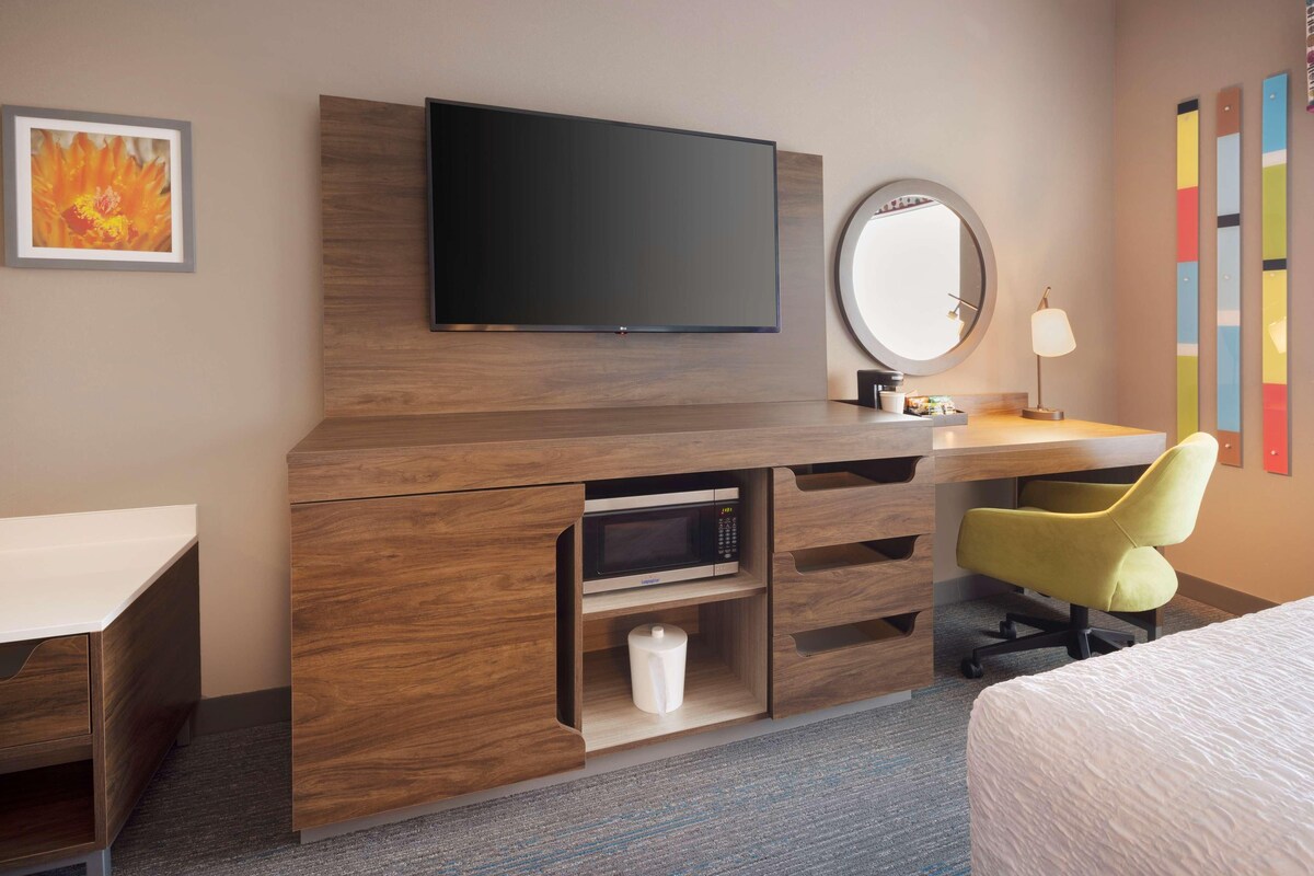 A hotel room with a wooden desk, a flat-screen TV, a round mirror, a yellow chair, and a bed.