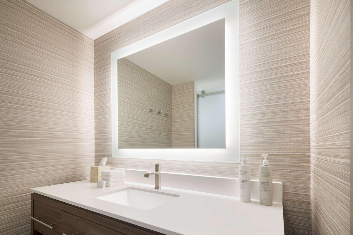 Un baño moderno con un espejo grande, un lavabo blanco y dos botellas de champú.