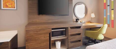 A hotel room with a wooden desk, a flat-screen TV, a mirror, a lamp, and a bed.