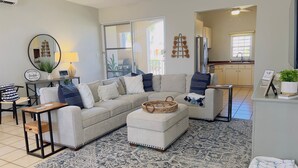 A modern living room with a beige sectional sofa, a coffee table, and a patterned rug. There is a kitchen area visible in the background.