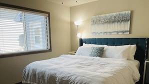 A neatly made bed with white linens, a dark blue upholstered headboard, and a single decorative pillow. A wall-mounted artwork is visible above the bed, and a floor lamp is positioned to the right. - Cozy 4King bed+3Bath near Downtown&Airport open concept Scenic backyard view (Sioux Falls)