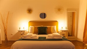 Un lit bien fait, avec du linge de lit blanc et un plaid vert, flanqué de deux tables de chevet avec des lampes, dans une pièce dotée d’un grand miroir et d’appliques murales décoratives.