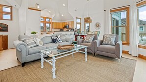 A spacious living room with a glass coffee table, two sofas, and a kitchen in the background. - Modern Beachhouse, Pool, 73 Steps to Beach, Deck Fully Loaded, Arcade (Nags Head)