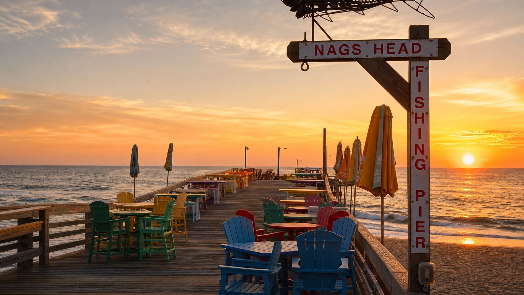 Eine Holzstege mit Tischen und Stühlen, ein Schild mit der Aufschrift 'NAGS HEAD FISHING PIER' und der Ozean im Hintergrund.