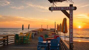 A wooden pier with tables and chairs, a sign indicating 'NAGS HEAD FISHING PIER', and the ocean in the background.