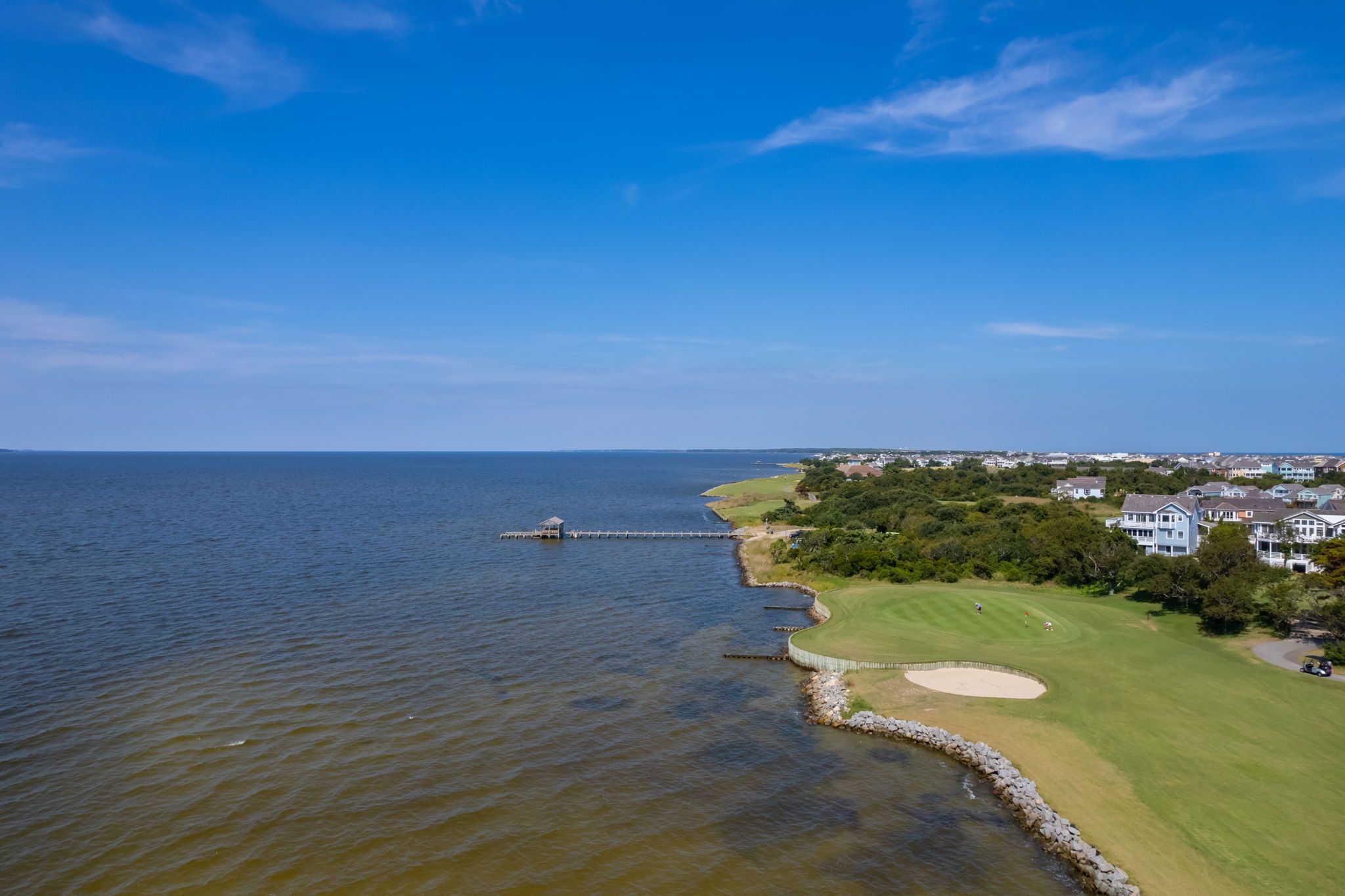 An aerial view of a coastal area with a golf course, a pier, and residential buildings.