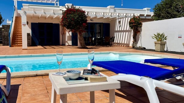 A pool area with lounge chairs and a table, a staircase leading to a building, and a large tree with red flowers. - Beautiful Villa with a private heated pool and roof terrace. (Playa Blanca)