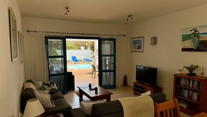 A living room with a sofa, TV, and bookshelf, a dining table, and a view of an outdoor pool area. - Beautiful Villa with a private heated pool and roof terrace. (Playa Blanca)