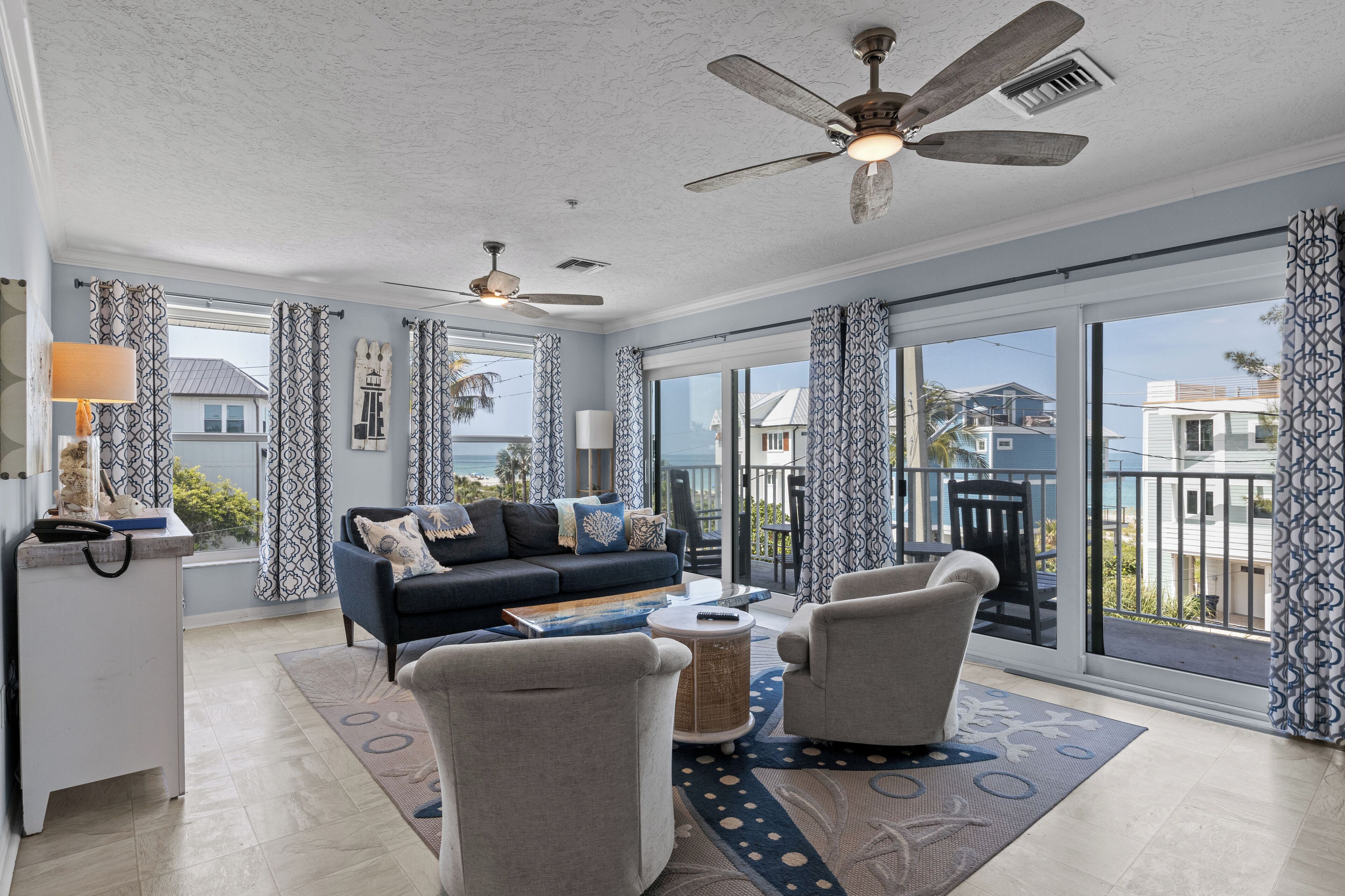 A spacious living room with a sofa, armchairs, and a coffee table. There are ceiling fans, a desk with a lamp, and large windows offering a view of the ocean and neighboring houses.